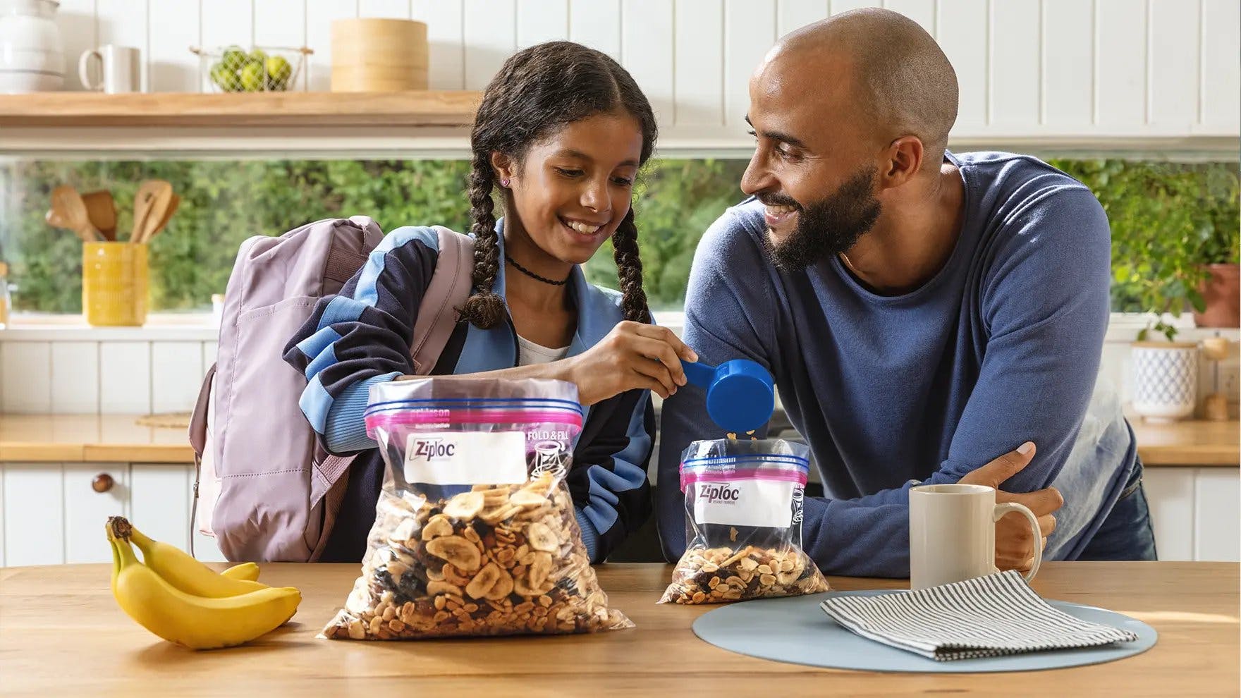 Father and daughter smiling and scooping snack mix into Ziploc quart-sized storage bag.